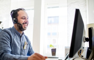 En leende person som sitter med ett headset framför en datorskärm