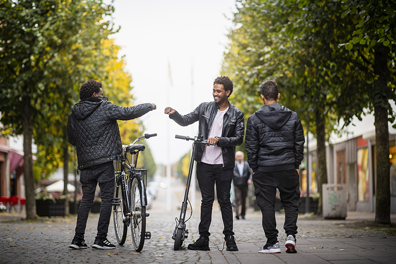 Tre unga män möts på gatan. En har en cykel och en har en elsparkcykel.