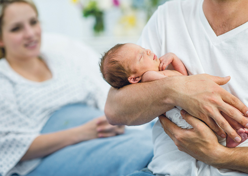 Nyfödd i armarna på sin pappa. Mamma i bakgrunden i sjukhussäng.