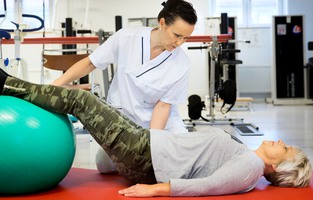 En sjukgymnast hjälper en kvinna  som ligger på rygg och har fötterna på en pilatesboll.