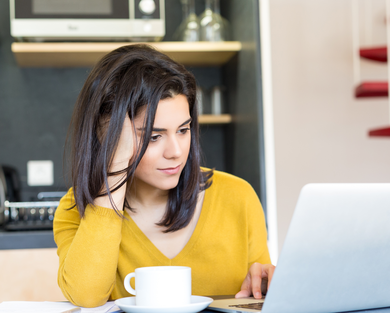 Kvinna sitter i ett kök med en dator och en kaffekopp.