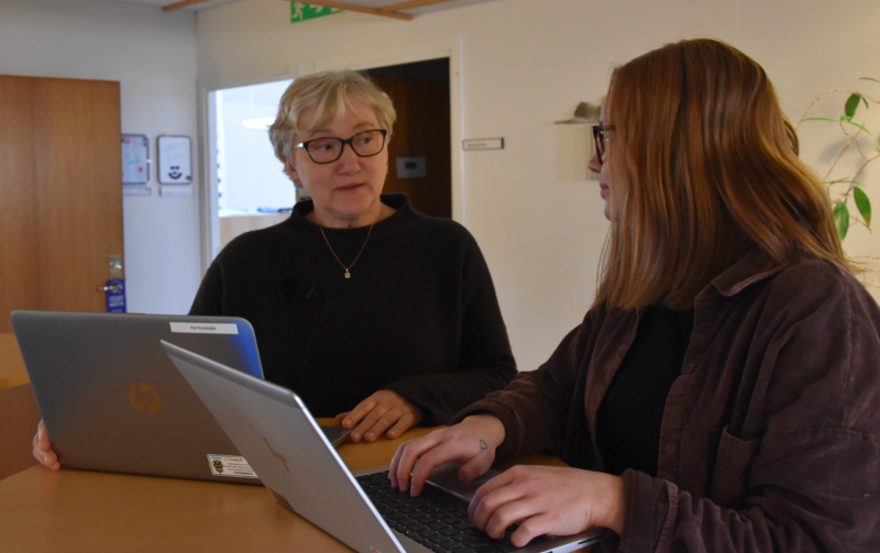 Kristina Luhr står med en kollega vid ett bord i en korridor. Båda har en laptop framför sig.