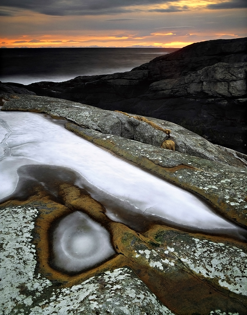 Fotografi av is på klippor vid hav. Solnedgång. Mörk himmel.