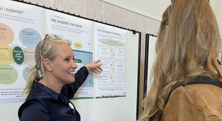 SamKrafts utvecklingsledare Camilla Pettersson berättar för en besökare på mässan. Hon pekar på posters på mässväggen bakom dem.