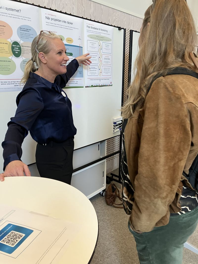 SamKrafts utvecklingsledare Camilla Pettersson berättar för en besökare på mässan. Hon pekar på posters på mässväggen bakom dem.