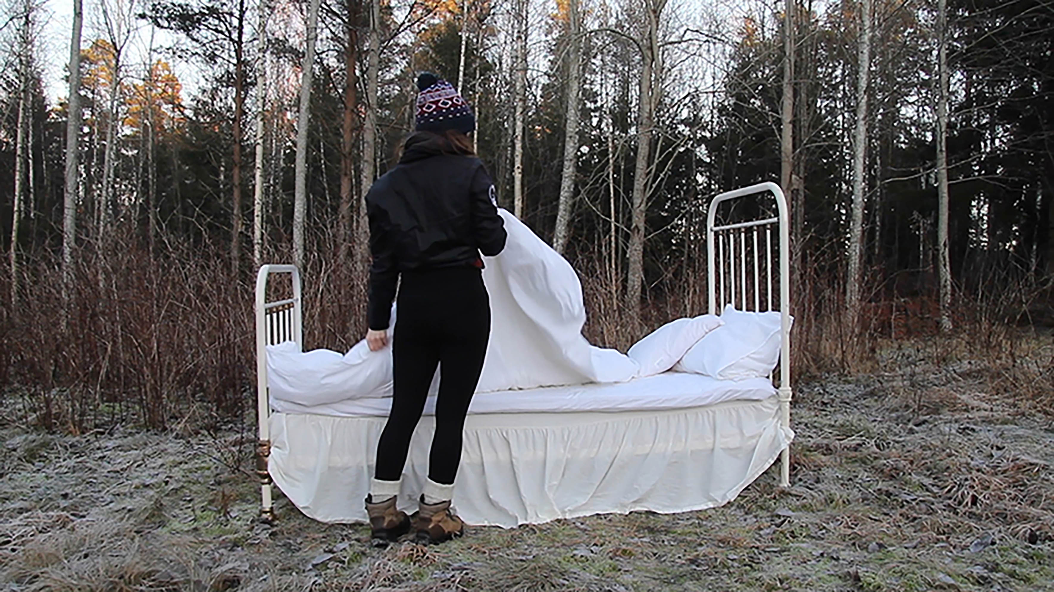 Fotografi av kvinna som står utomhus vid en gammal, vit säng med vita lakan.