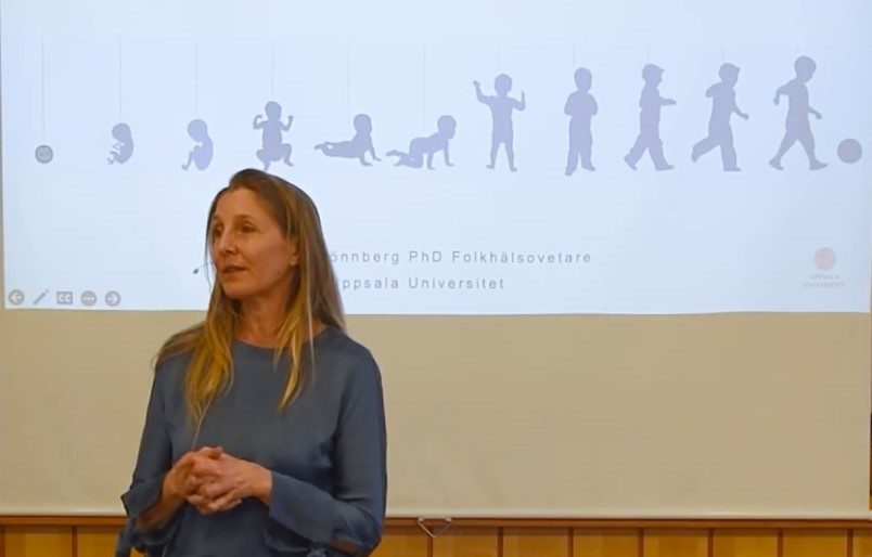 Gunilla Lönnberg, en kvinna med långt blont hår. Hon har en blå blus. Bakom henne finns en skärm med illustrationer av barn från foster till en tvååring som springer efter en boll.