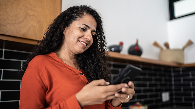 Mörkhårig kvinna står i ett kök och tittar på en mobiltelefon som hon håller i händerna.