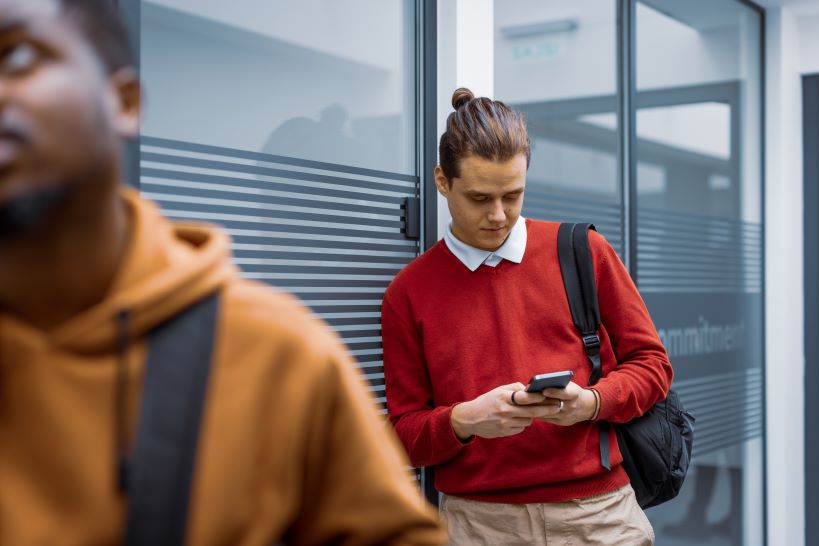 En kille med röd tröja som tittar ner i mobilen, han har skjorta under tröjan. Runt 20 år 
