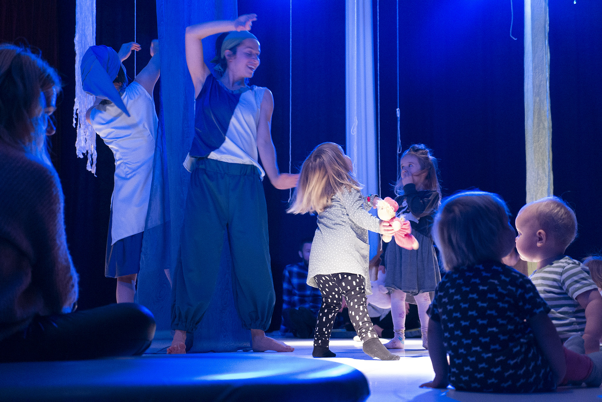 Två dansare rör sig tillsammans med små barn som sitter och står på ett scengolv i blåaktigt ljus