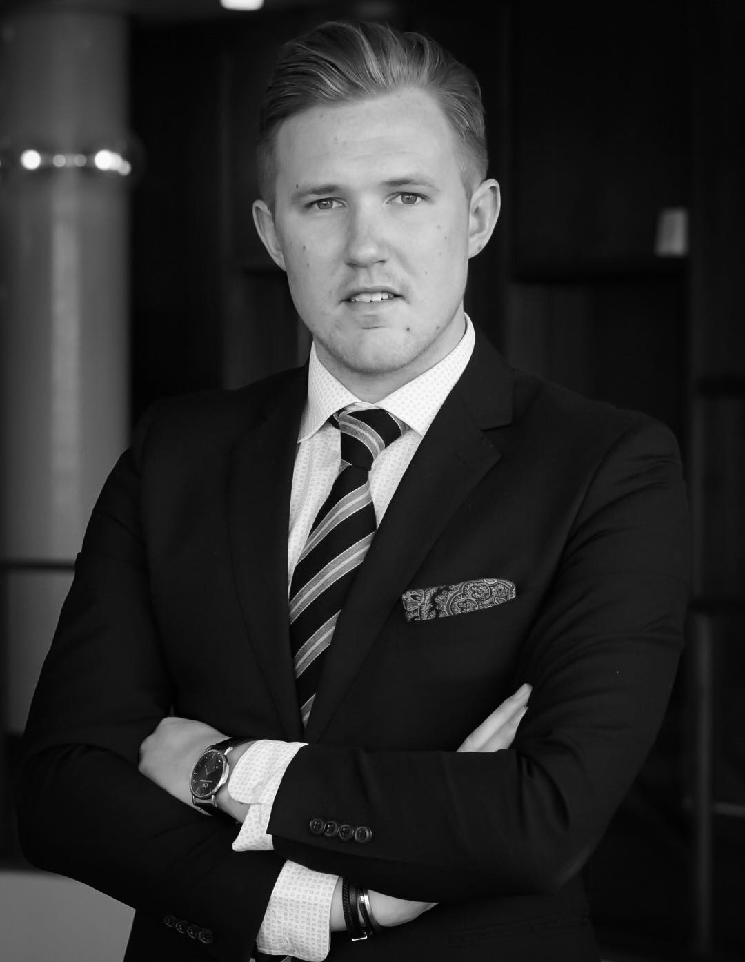 I en svartvit bild står en man i kostym med händerna i kors och tittar in i bild.