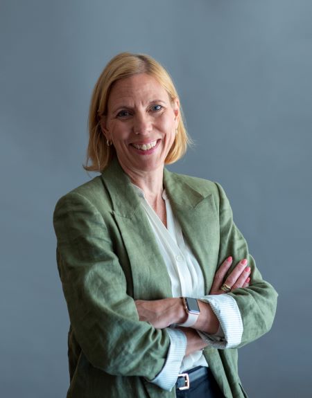En blond kvinna i grön kavaj står leende med korslagda armar.