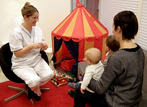 Tandvårdspersonal i vita kläder sitter och pratar med en förälder som har två barn i sin famn.
